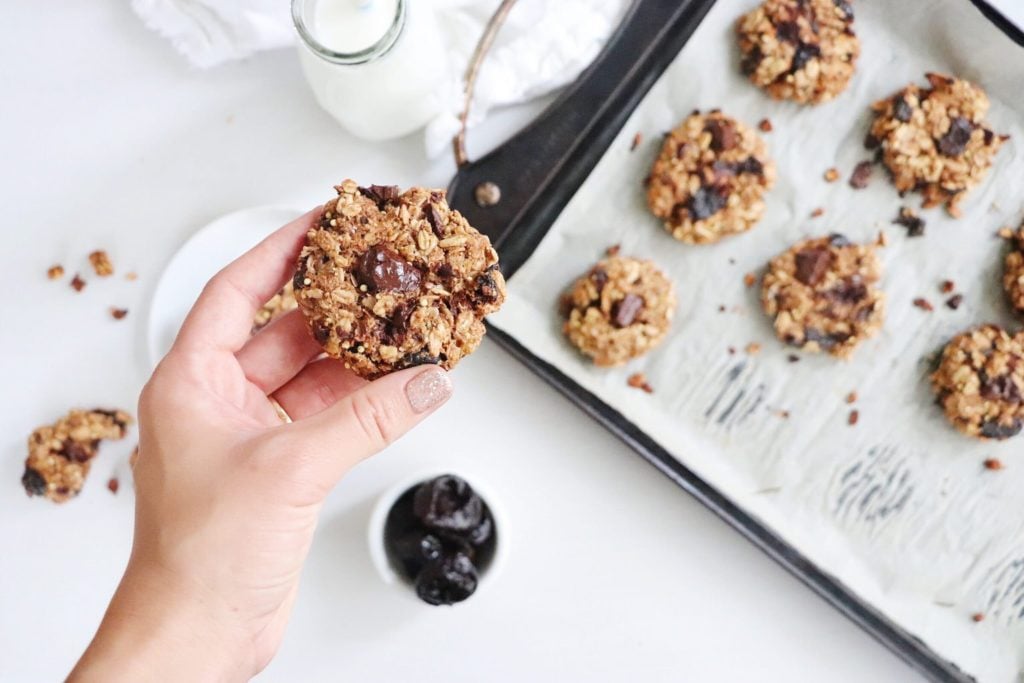Lindsay Pleskot holding a One Bowl Breakfast Power Cookie in her hand over top of a white food photography board that has a cookie sheet lined with parchment paper and power cookies. Ingredients include oats, prunes, chocolate chips.