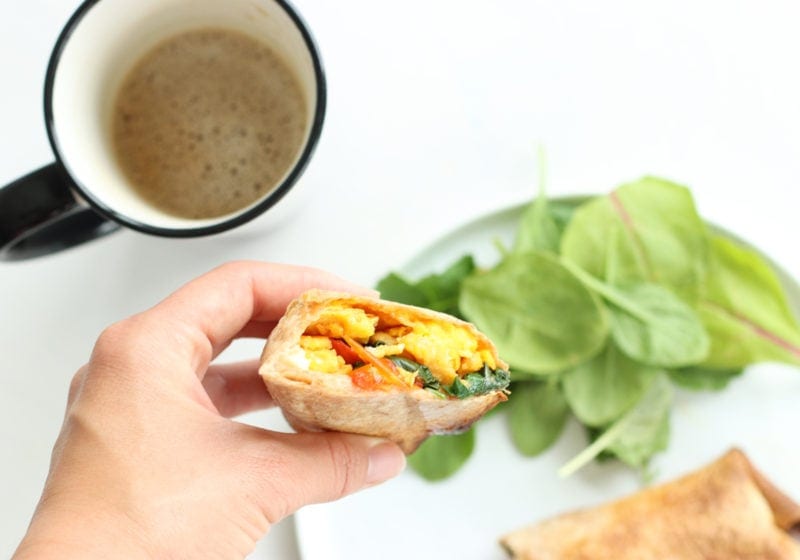 Lindsay Pleskot holding a healthy prep ahead breakfast burrito wrap. Ingredients include: eggs, whole grain wraps, olive oil, spinach, onion, bell pepper, feta, salt and pepper.