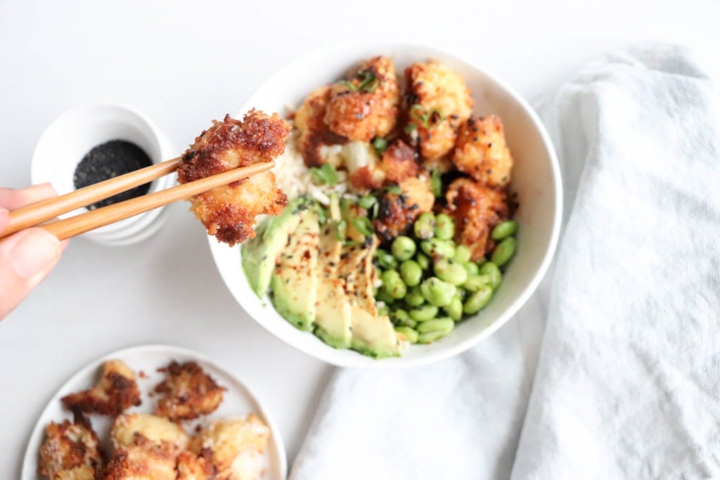 Crispy cauliflower bowl over a white surface with a kitchen towel beside it. Ingredients include cauliflower, bread crumbs, edamame, avocado, rice.