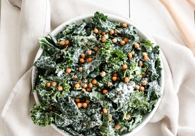 Chickpea kale caesar salad on a white plate over a neutral kitchen dish. Ingredients include: kale, chickpeas, capers, Greek yogurt, and garlic.