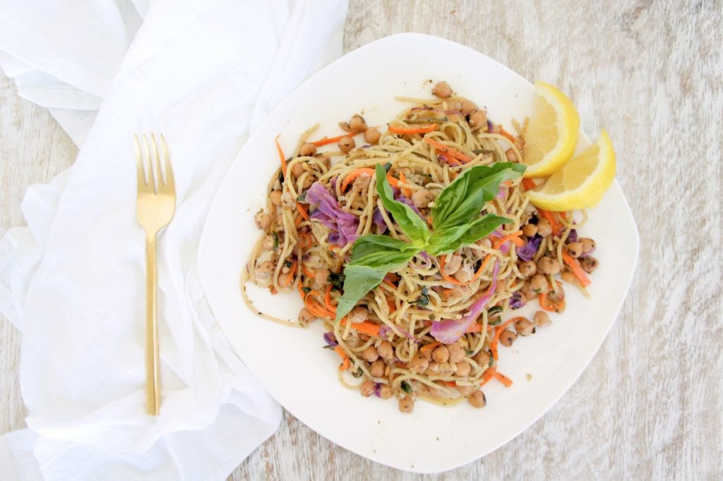 Chickpea pasta on a white plate with lemon wedges. Ingredients include: spaghetti noodles, carrot, zucchini, purple cabbage, chickpeas, garlic, lemon.