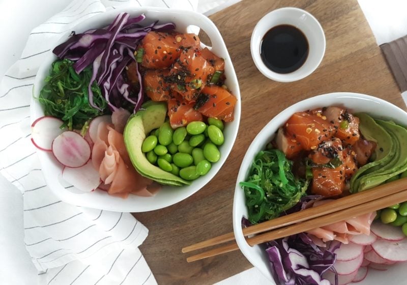 Two white poke bowls on a wooden food board with a striped kitchen towel beside it. Ingredients include: salmon, edmame, avocado, cabbage, wakame salad, pickled ginger, black sesame seeds.