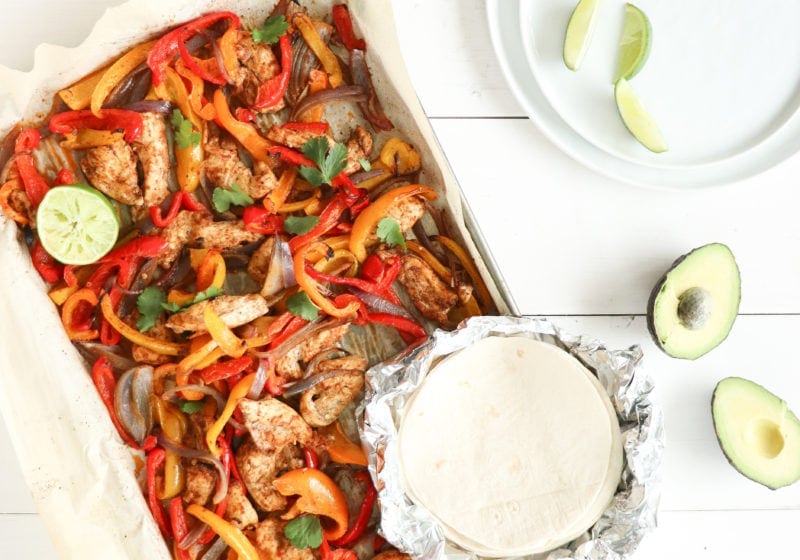 Sheet pan fajitas over a white food board with an avocado sliced in half beside it. Ingredients include bell peppers, chicken, spices, lime.