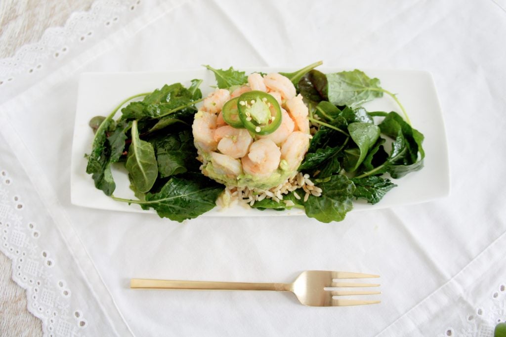 Shrimp stack over a white serving plate. Ingredient sinclude leafy greens, brown rice, avocado, shrimp.