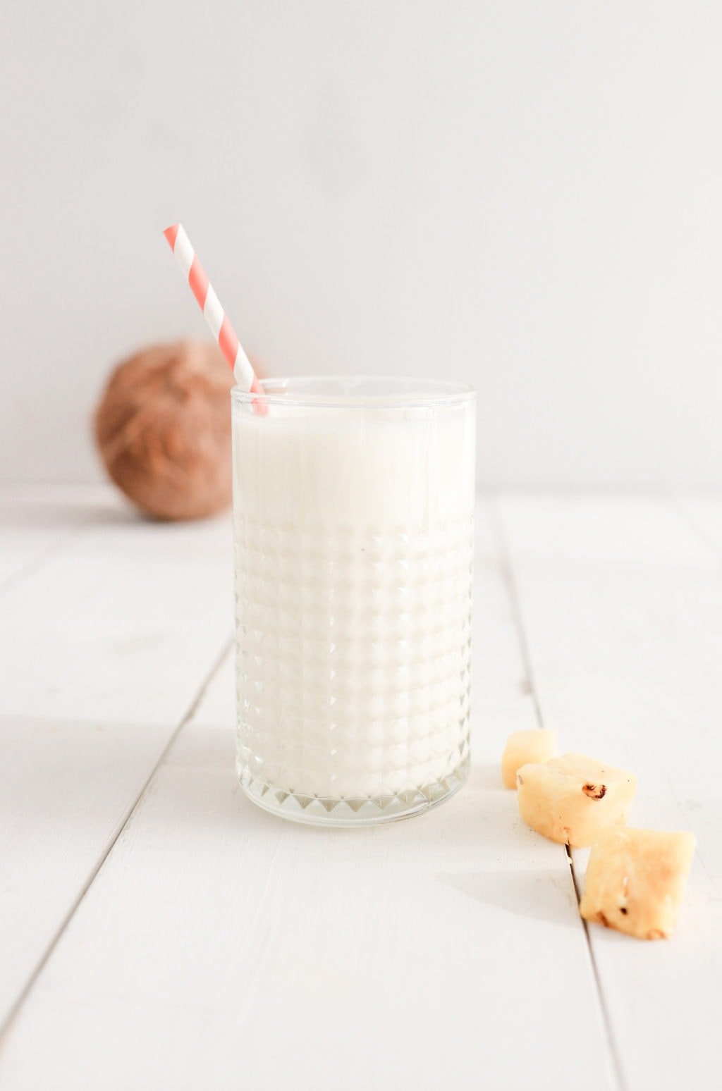 A pina colada smoothie in a glass with a pink and white striped straw on a white surface with pineapple chunks beside it and a coconut in the background. Ingredients include coconut milk, pineapple, banana, and plain Greek yogurt