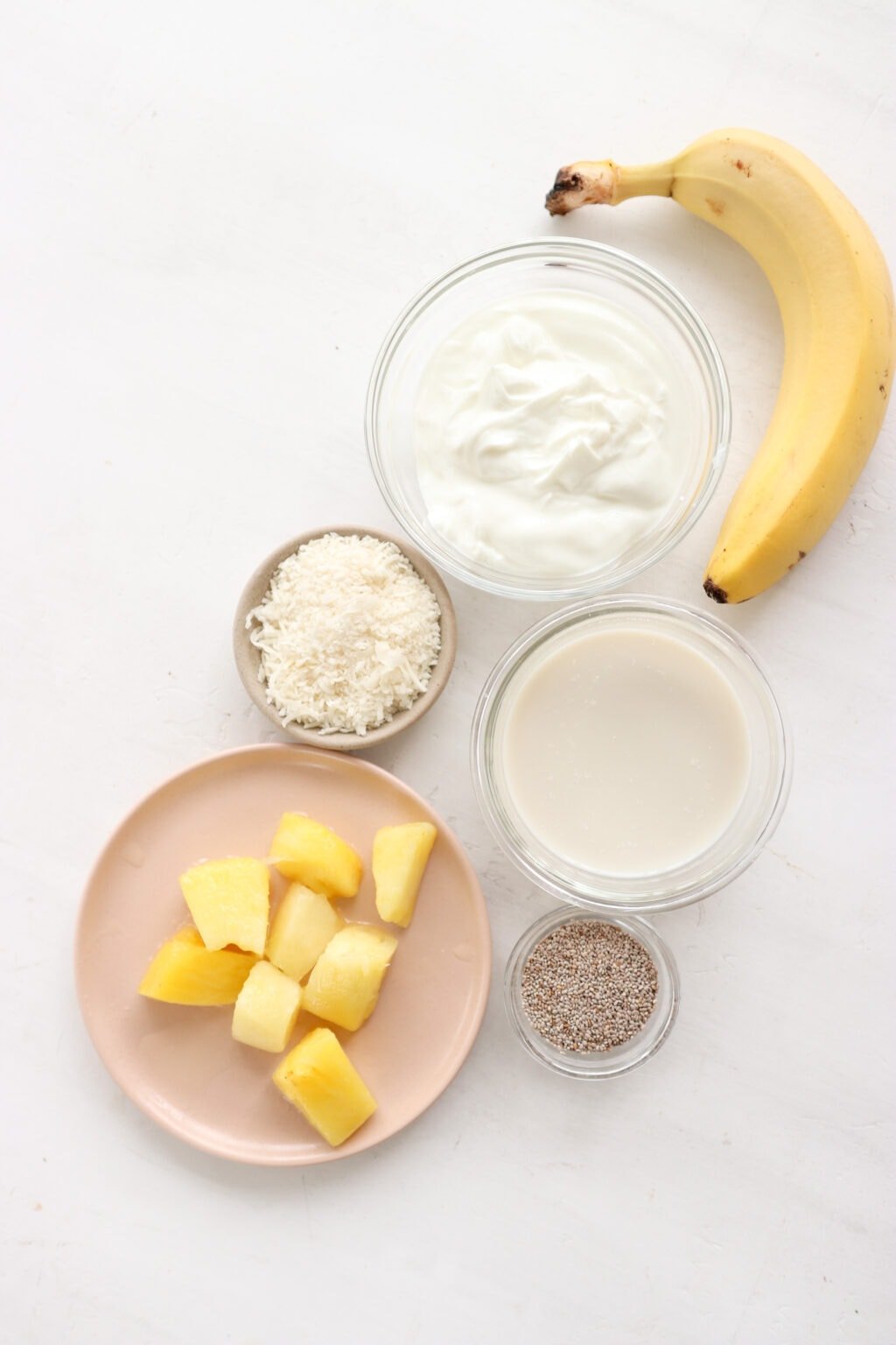 Recipe ingredients laid out on a counter including a banana, greek yogurt, coconut, coconut milk, chia seeds, and pineapple.