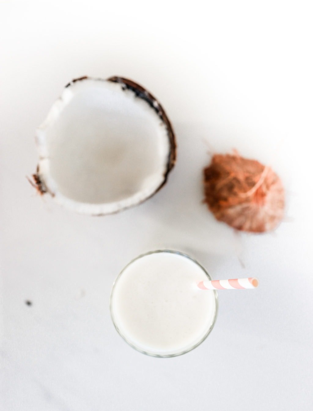 A pina colada smoothie in a glass with a pink and white striped straw on a white surface with a broken coconut beside it. Ingredients include coconut milk, pineapple, banana, and plain Greek yogurt
