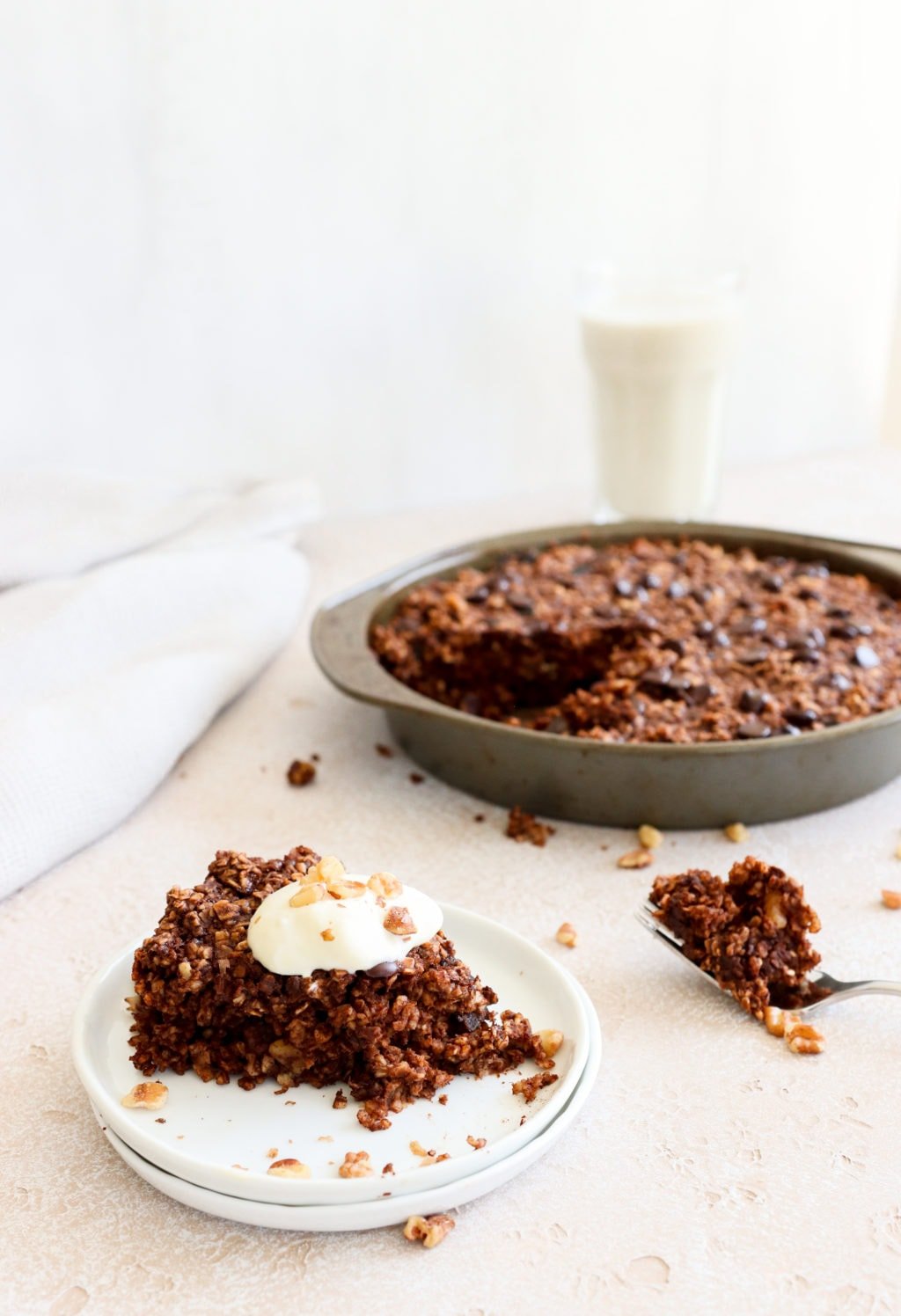 A slice of chocolate baked oatmeal on two white plates with the pan of chocolate baked oatmeal behind. Ingredients include: oats, banana, egg, coconut oil, maple syrup, cinnamon, prunes, walnuts.