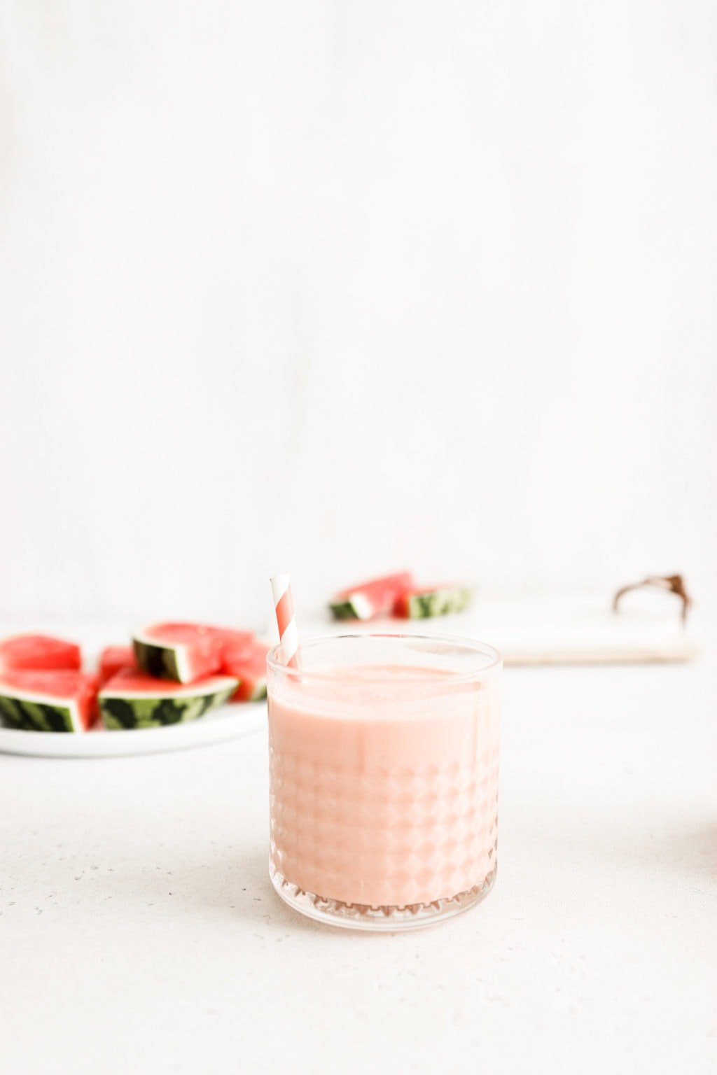 A watermelon banana smoothie in a glass cup with a striped pink and white straw. The smoothie is on a neutral surface with slices of watermelon faded in the background. Ingredients include: watermelon, banana, Greek yogurt, lime.