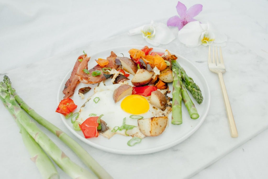 A marble slab with a plate of Healthy Sheet Pan Breakfast Bake on it and 3 sprigs of asparagus beside it on the left and a gold fork and flowers on the right. Ingredients include: bacon, eggs, bell peppers, potatoes, asparagus.
