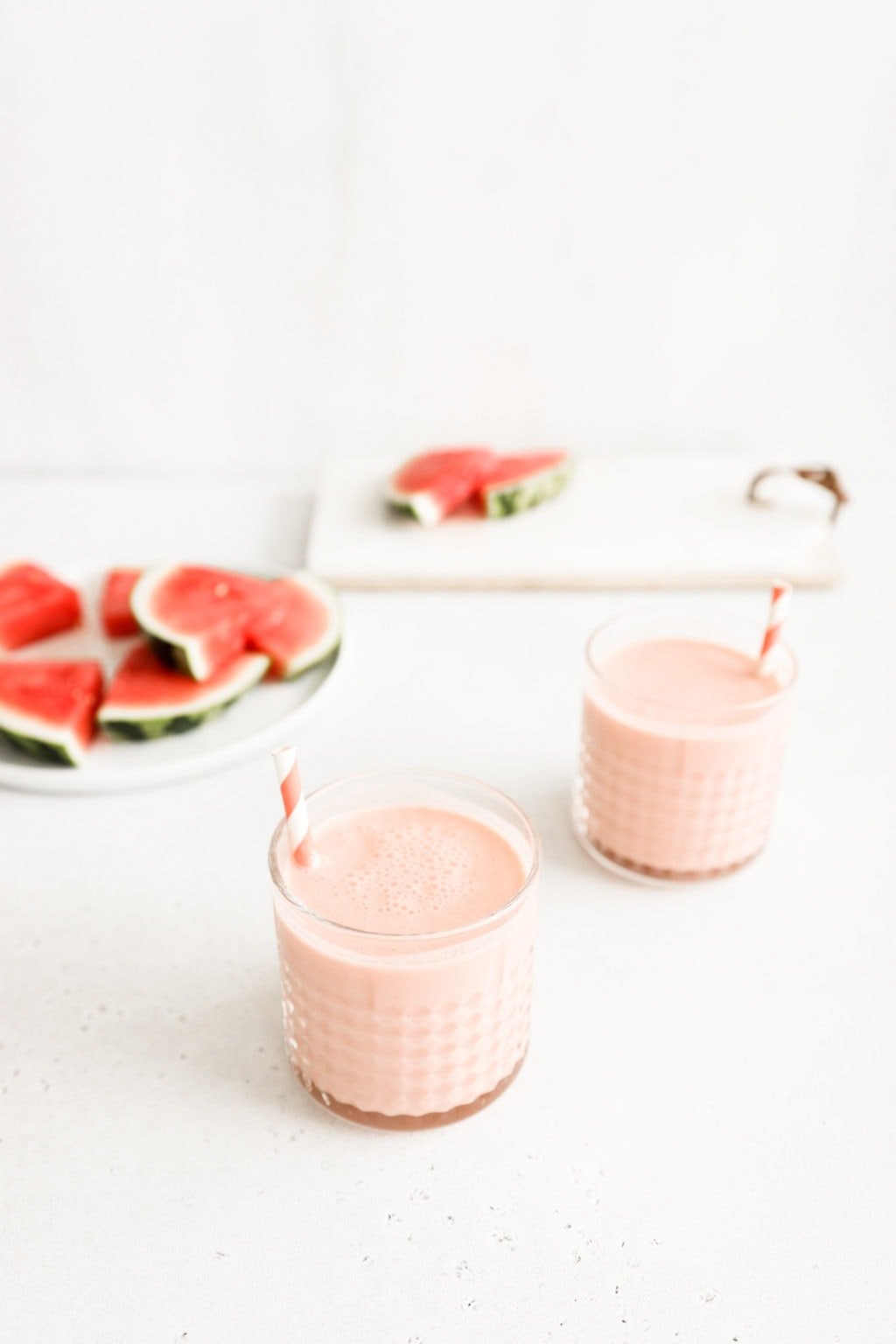 Two watermelon banana smoothies in a glass cup with a striped pink and white straw. The smoothies are on a neutral surface with slices of watermelon beside them. Ingredients include: watermelon, banana, Greek yogurt, lime.