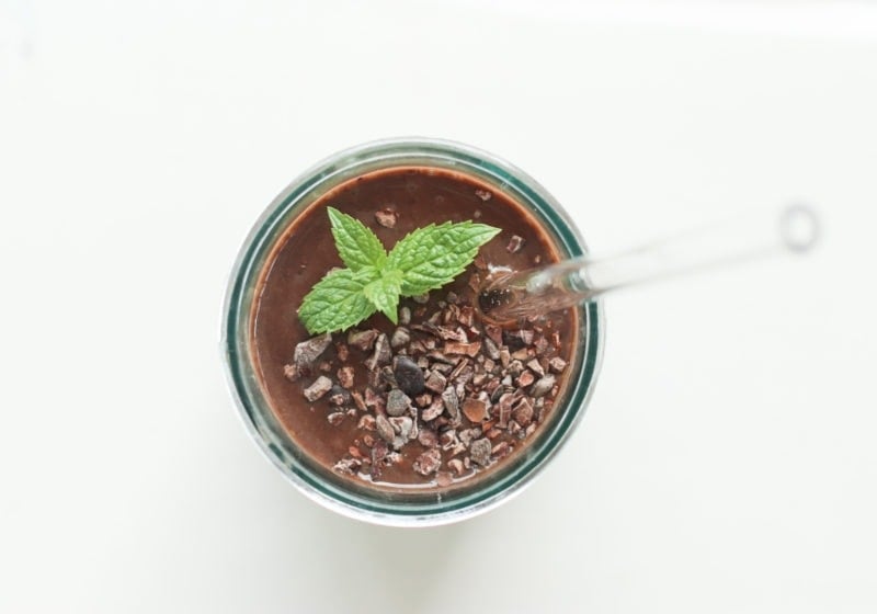 Mint Cocolate Black Bean smoothie over a white surface in a glass jar with a clear straw. Garnished with mint and cacao nibs.