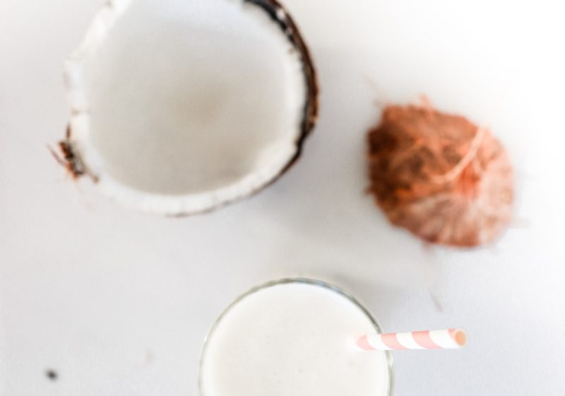 A pina colada smoothie in a glass with a pink and white striped straw on a white surface with a broken coconut beside it. Ingredients include coconut milk, pineapple, banana, and plain Greek yogurt