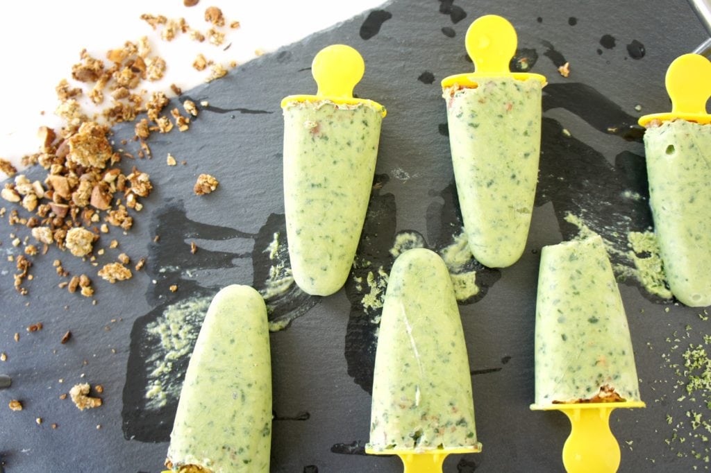 Key Lime Kefir Smoothie Pops on a black food board with granola spread out beside it. Ingredients include spinach, avocado and kefir.