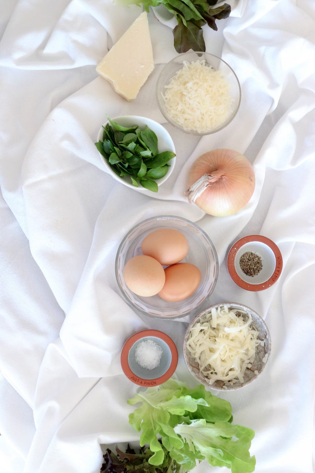 Ingredients for muffin tin frattatas are laid out on a white wrinkled tablecloth and include inspired greens lettuce, parmesan cheese, basil, onion, eggs, salt and pepper