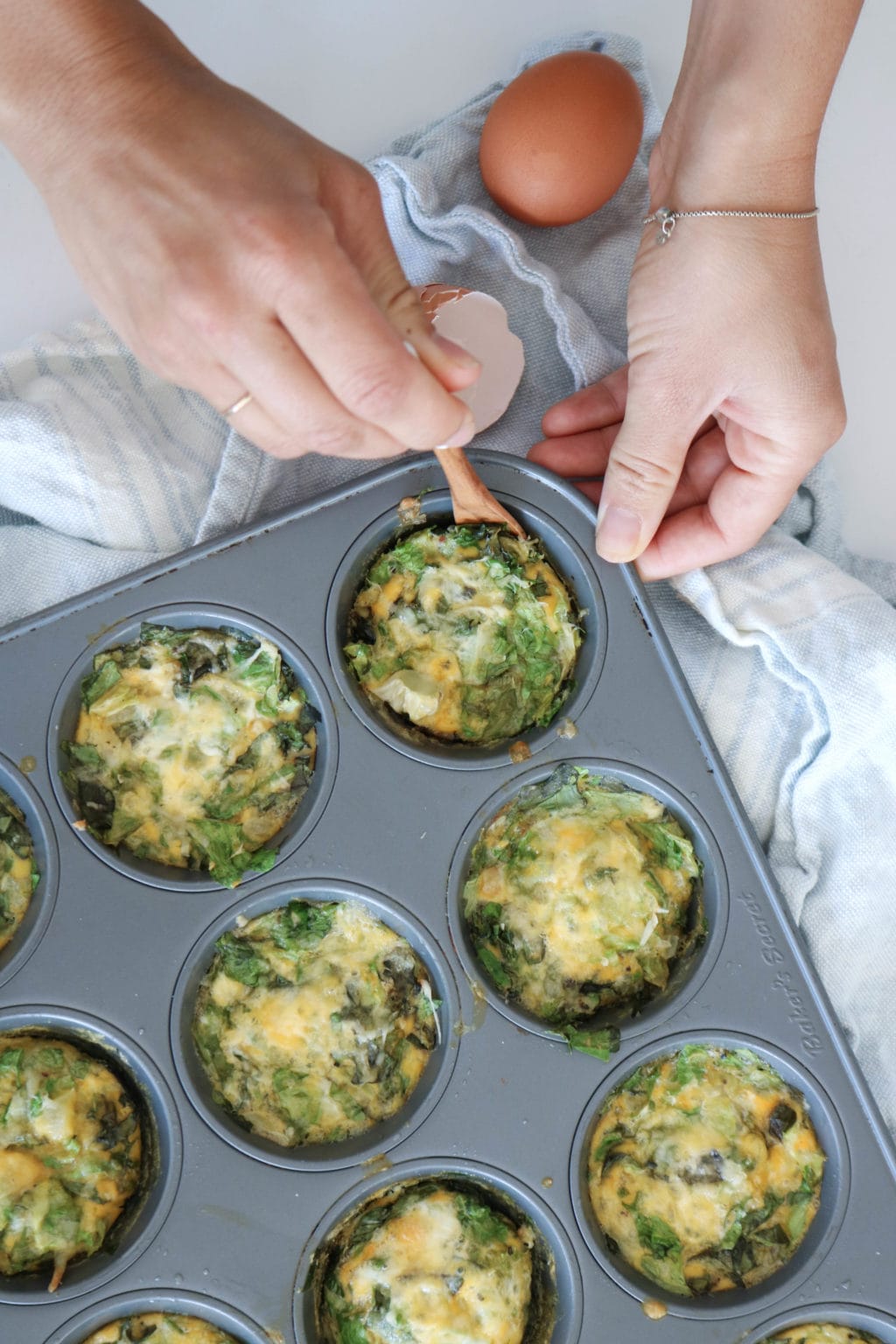 Muffin tin frittatas with eggs cheese and mushrooms are in a muffin tinlaid over a blue tea towel. Cracked egg shells are on the side and a woman's hand is scooping one of the frittatas out with a small wooden spoon