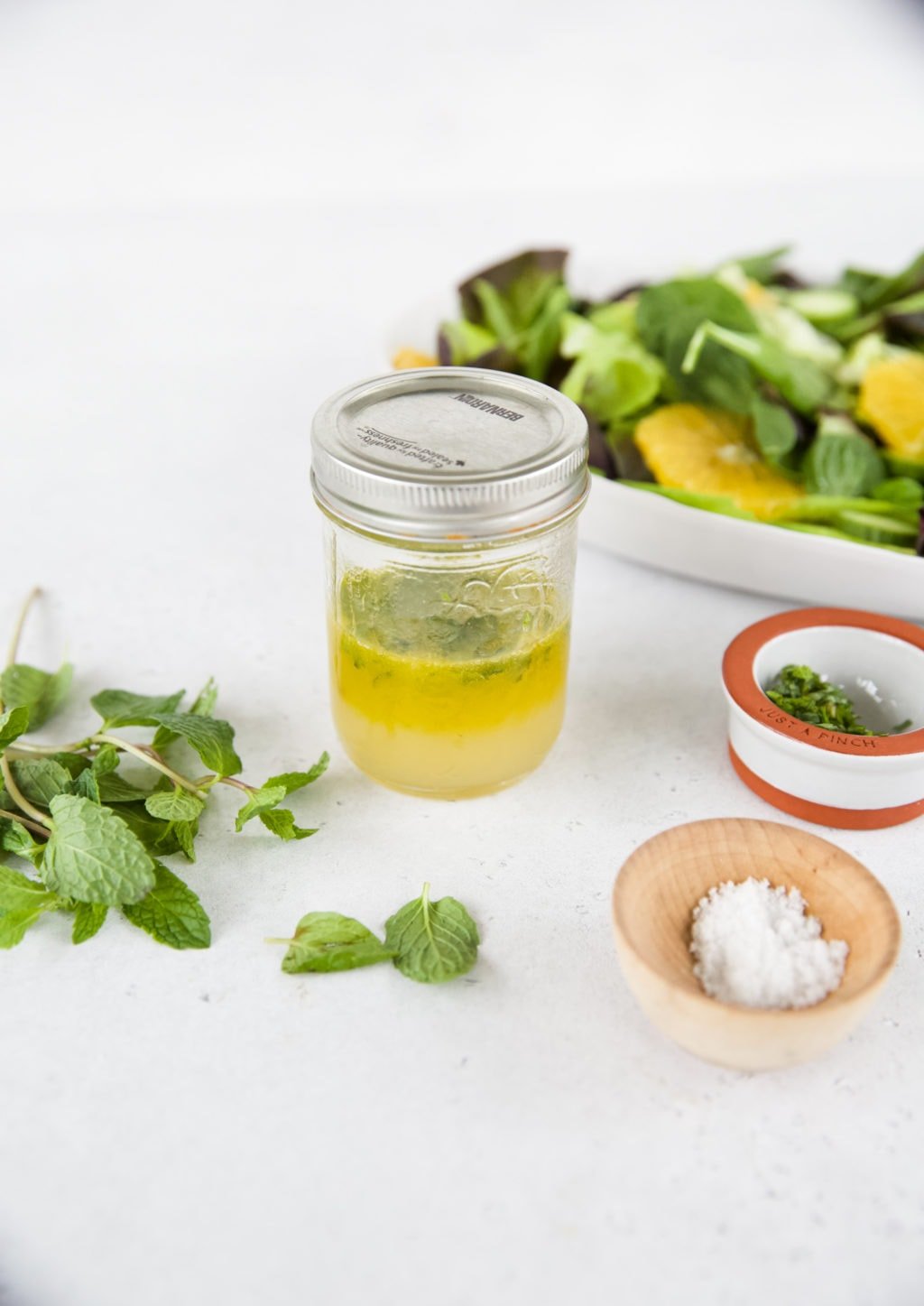 A small, 8 ounce mason jar is filled halfway with an olive oil based salad dressing. It sits on top of a white counter top. Mint leaves, a small bowl of salt, and a salad surround the jar.