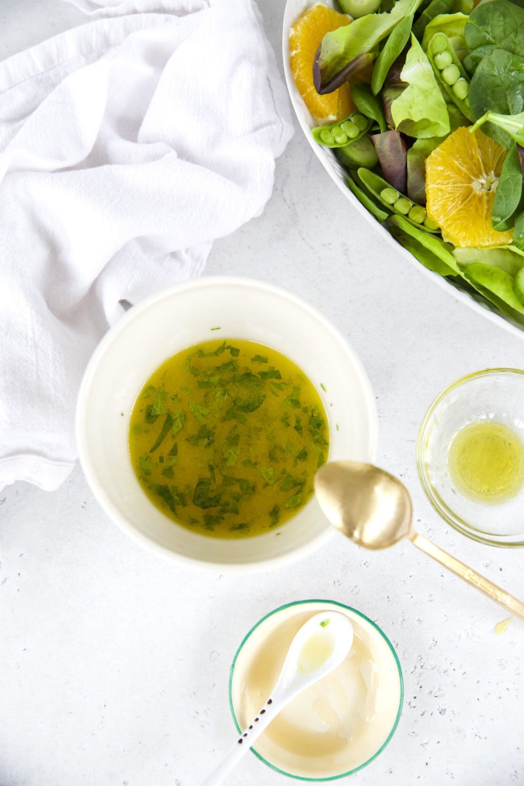 Honey, lime, and chopped mint salad dressing, sitting in a white bowl on top of a white counter with a gold spoon. Smaller bowls surround the dressing bowl, with a green salad in the top righthand corner. 