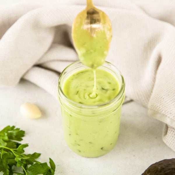 Avocado green goddess dressing dripping from a gold spoon into a full mason jar of the dressing