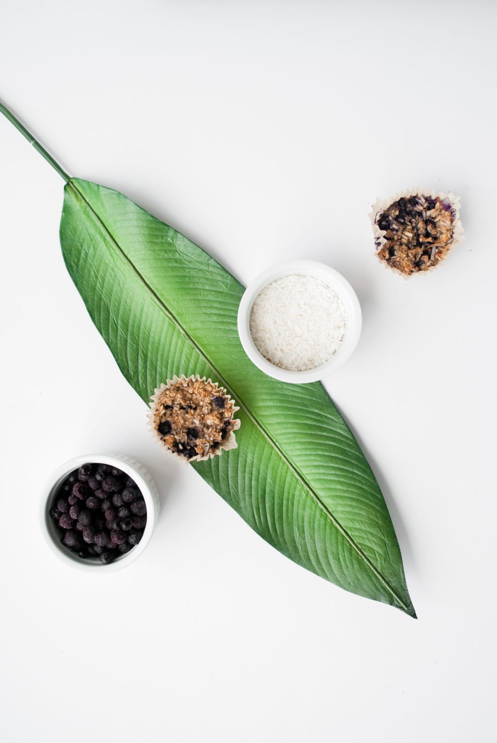 Bird's-eye view of two baked banana oatmeal cups between a bowl of blueberries and a bowl of shredded coconut on top of a tropical leaf and white backdrop.