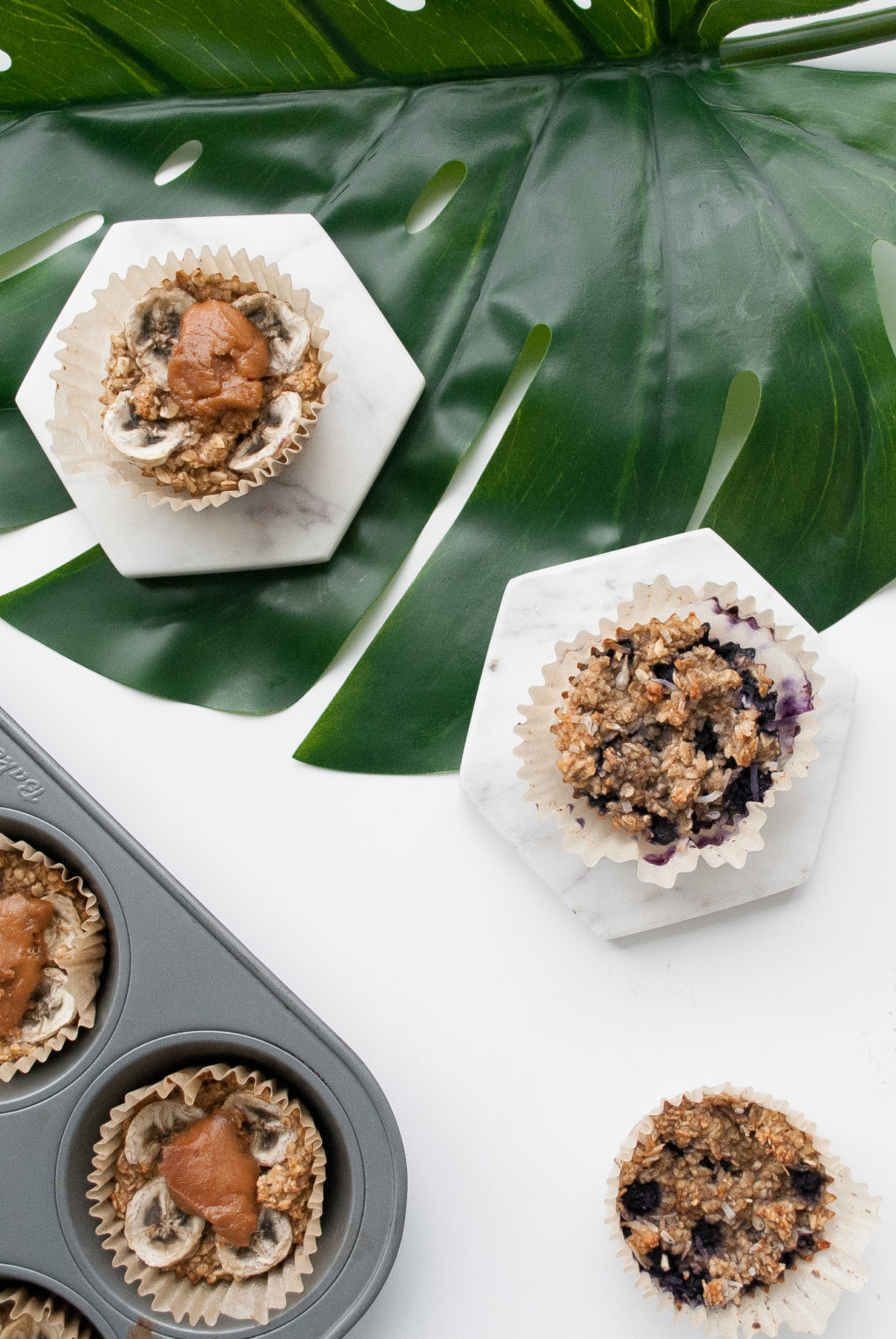 Bird's-eye view of two baked banana oatmeal cups next a muffin tray on top of a tropical leaf and a white backdrop.
