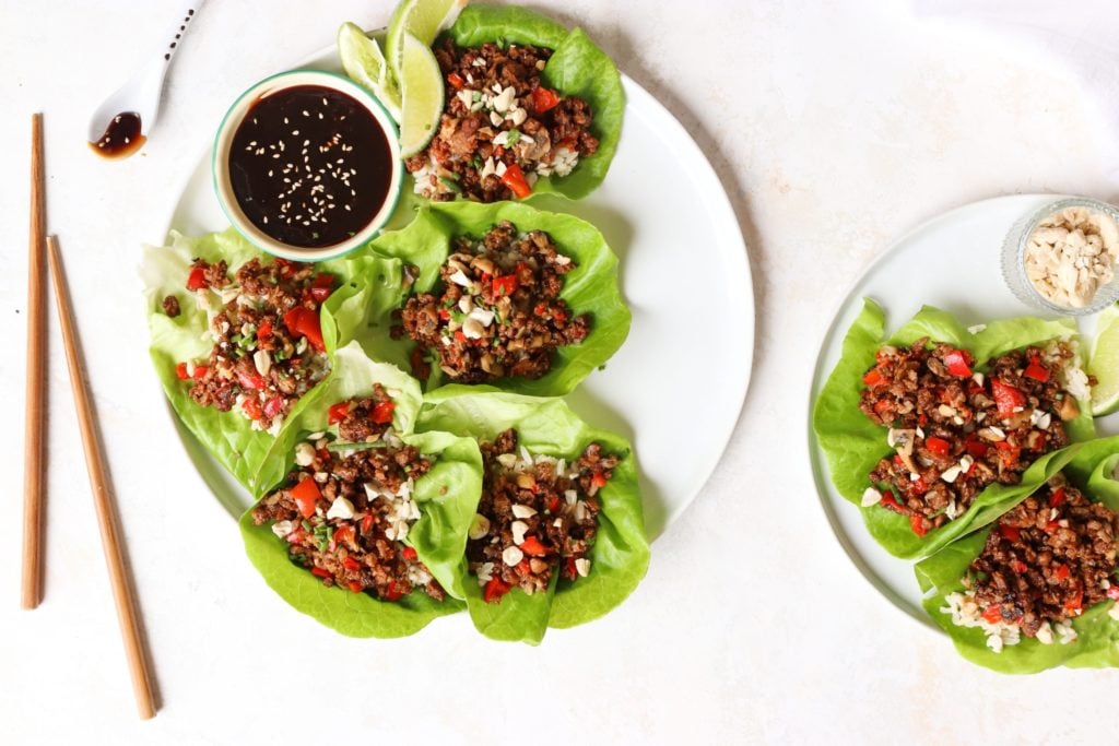 A photo of a white plate of 5 open face lettuce wraps and a small bowl of hoisin. At the top of the photo are chop sticks and a white spoon dripping with hoisin.