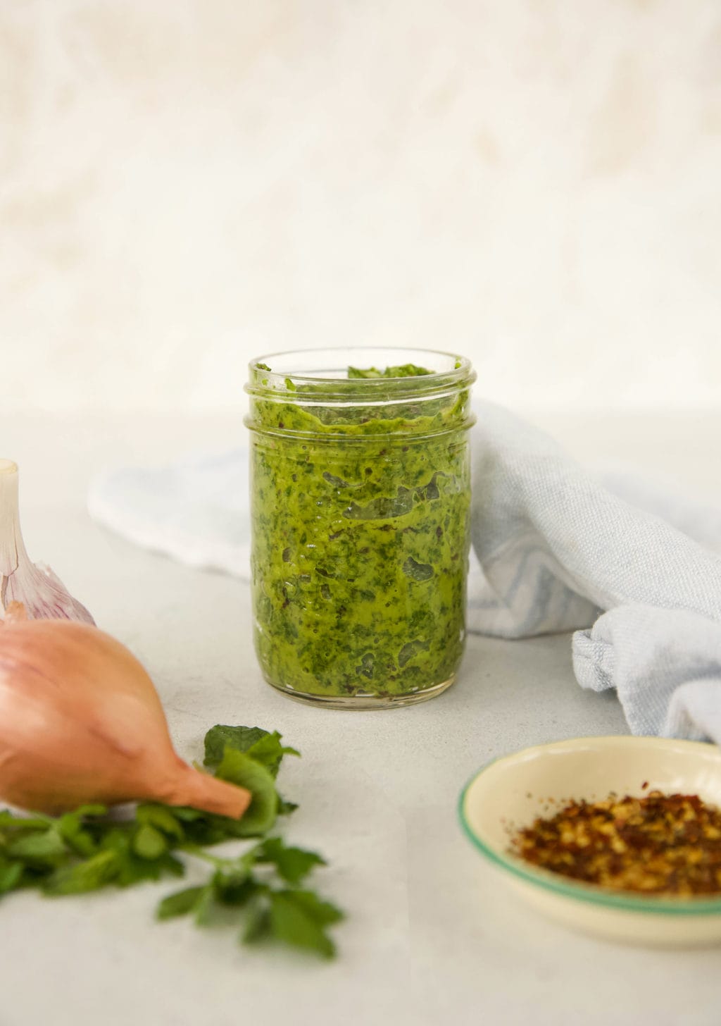 Cilatnro Chimichurri Sauce in a mason jar surrounded by ingredients (onion, red pepper flakes and basil)