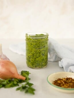 Cilatnro Chimichurri Sauce in a mason jar surrounded by ingredients (onion, red pepper flakes and basil)