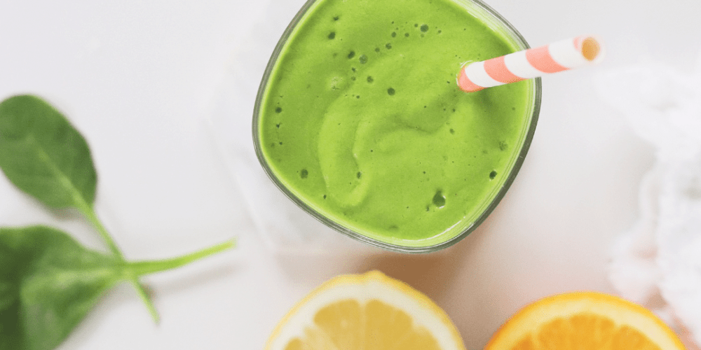 Green smoothie with red and white straw surrounded by oranges and kale