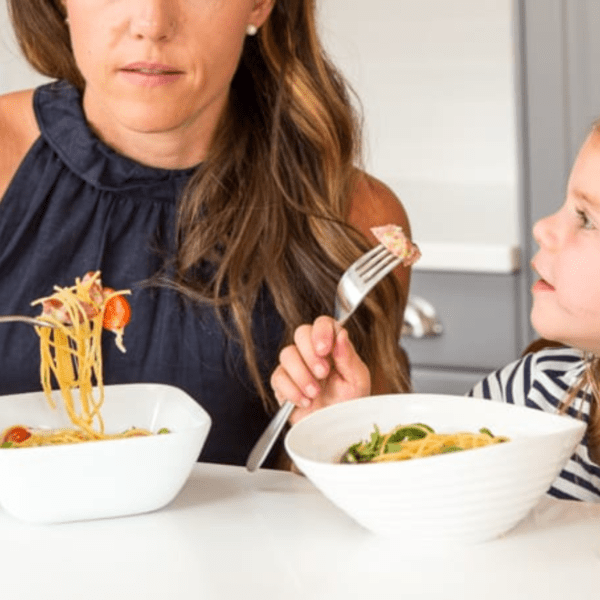 A woman in a nacy blue shirt is eating pasta and her daughter is looking up at her as she eats
