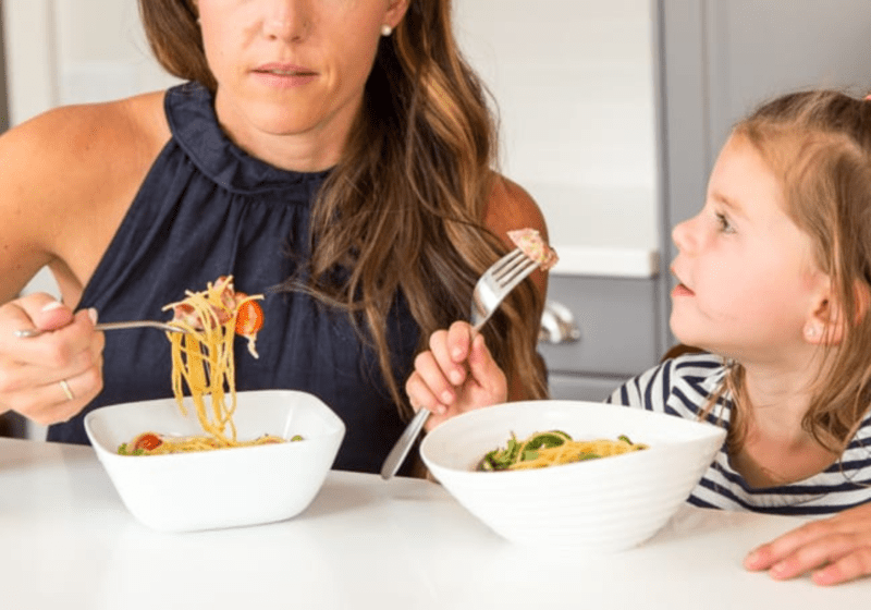 A woman in a nacy blue shirt is eating pasta and her daughter is looking up at her as she eats
