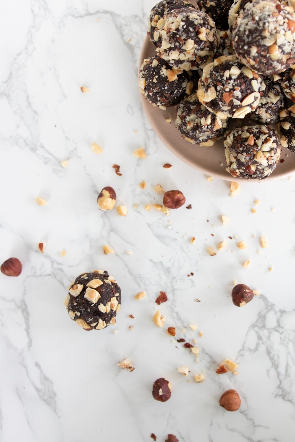 A single hazelnut energy ball is on a marble counter surrounded by crushed hazelnuts. A plate of energy bits is in the corner.