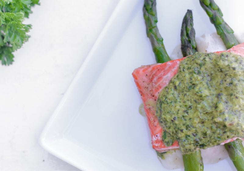 A white plate with baked salmon topped with chimichurri sauce sits on a plate of mashed potatoes and asparagus