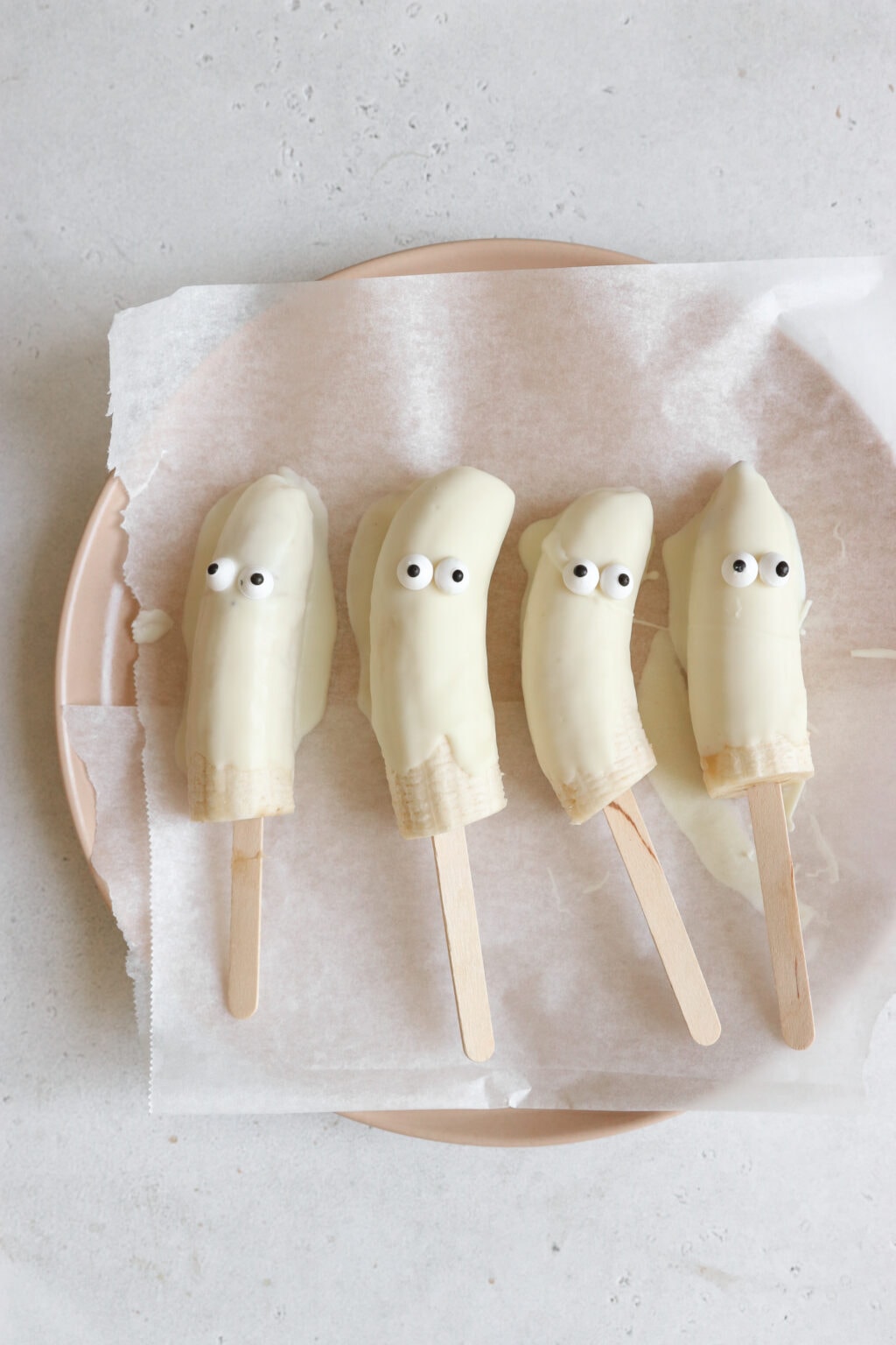 4 bananas are on a pink plate. The bananas are covered in white chocolate and have candy eyes and are on popsicle sticks. The bananas look like ghosts.