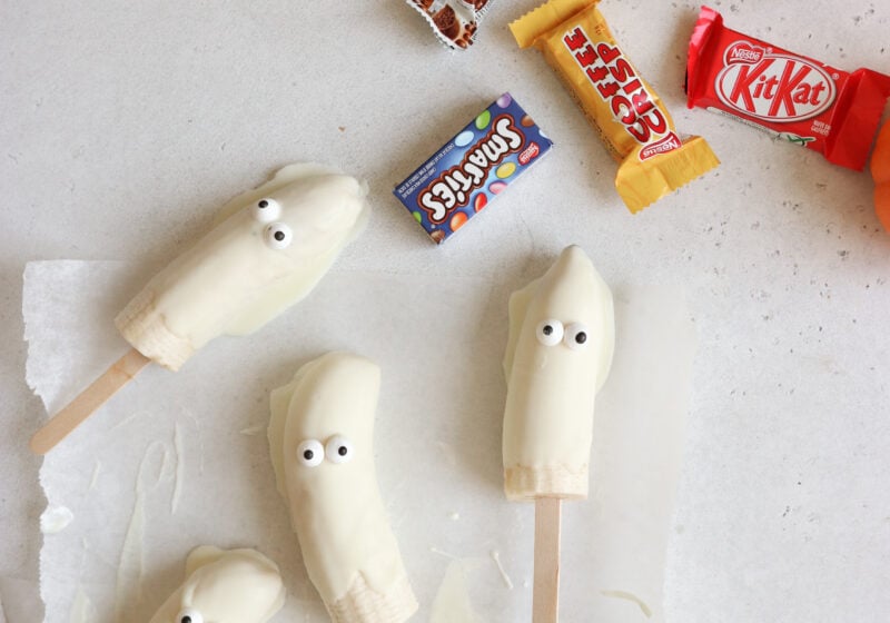 4 bananas covered in white chocolate with candy eyes and popsicle sticks are on parchment paper. There is an aero bar, smarties, coffee crisp and kitkat in the right hand corner. The candies are all Halloween size.