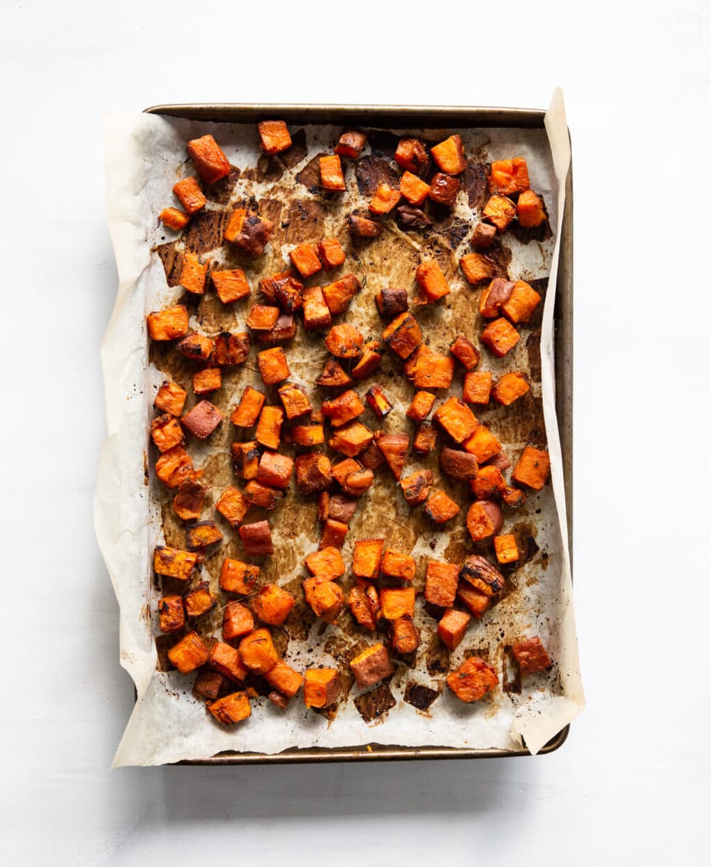 A baking sheet lined with parchment paper has golden brown, baked sweet potatoes on it. The sweet potatoes are cubed.