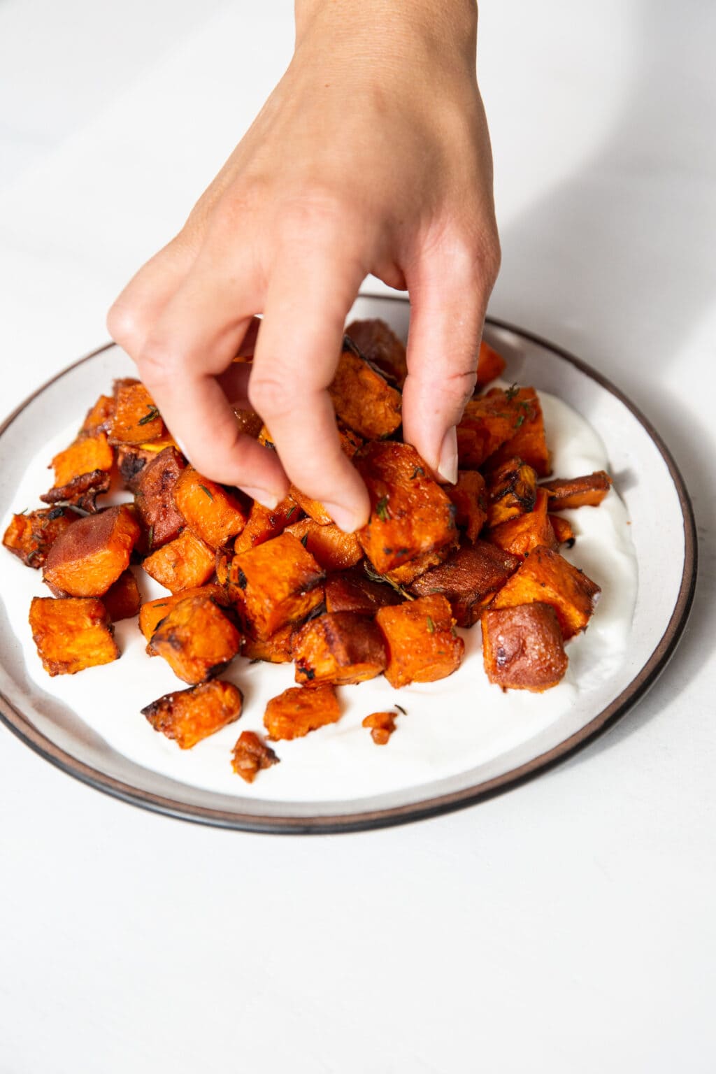 A hand is picking up a baked sweet potato cube on a white plate with a brown rim.