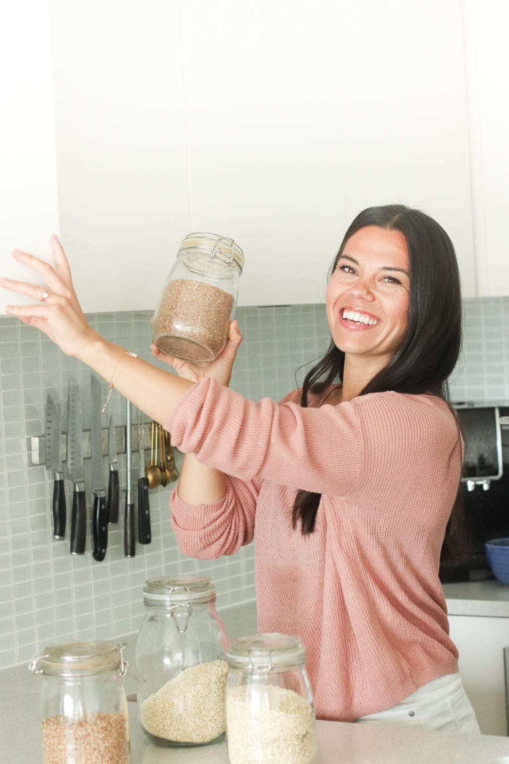 A girl with dark hair and a pink sweater is smiling at the camera. She is holding a jar of grains. In front of her is a jar of oats, rice and quinoa