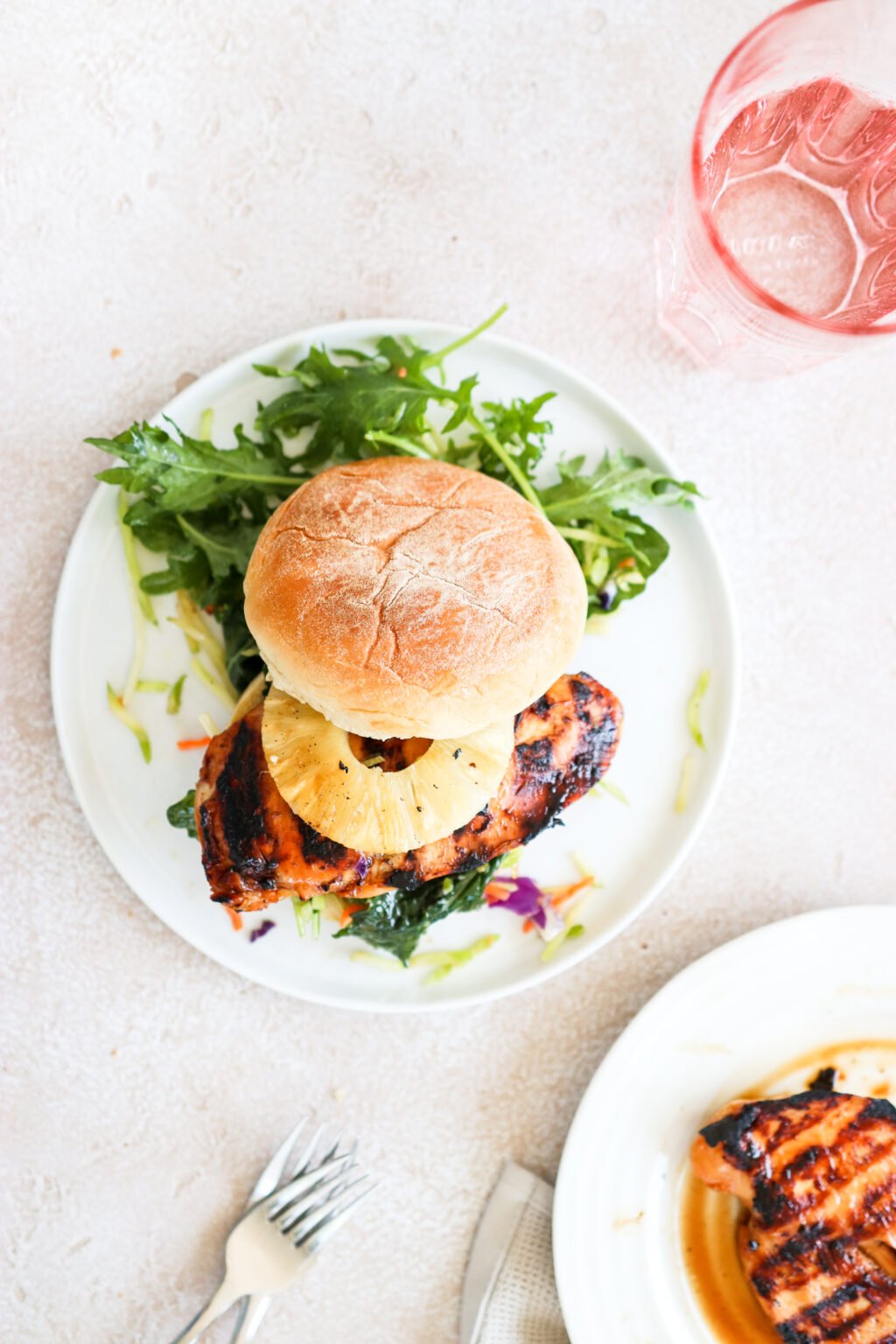 A grilled chicken burger topped with a pineapple ring is on a white plate and is surrounded by an arugula salad. 