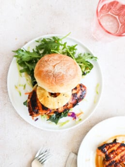 A grilled chicken burger topped with a pineapple ring is on a white plate and is surrounded by an arugula salad.