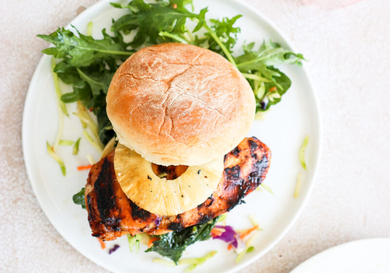 A grilled chicken burger topped with a pineapple ring is on a white plate and is surrounded by an arugula salad.