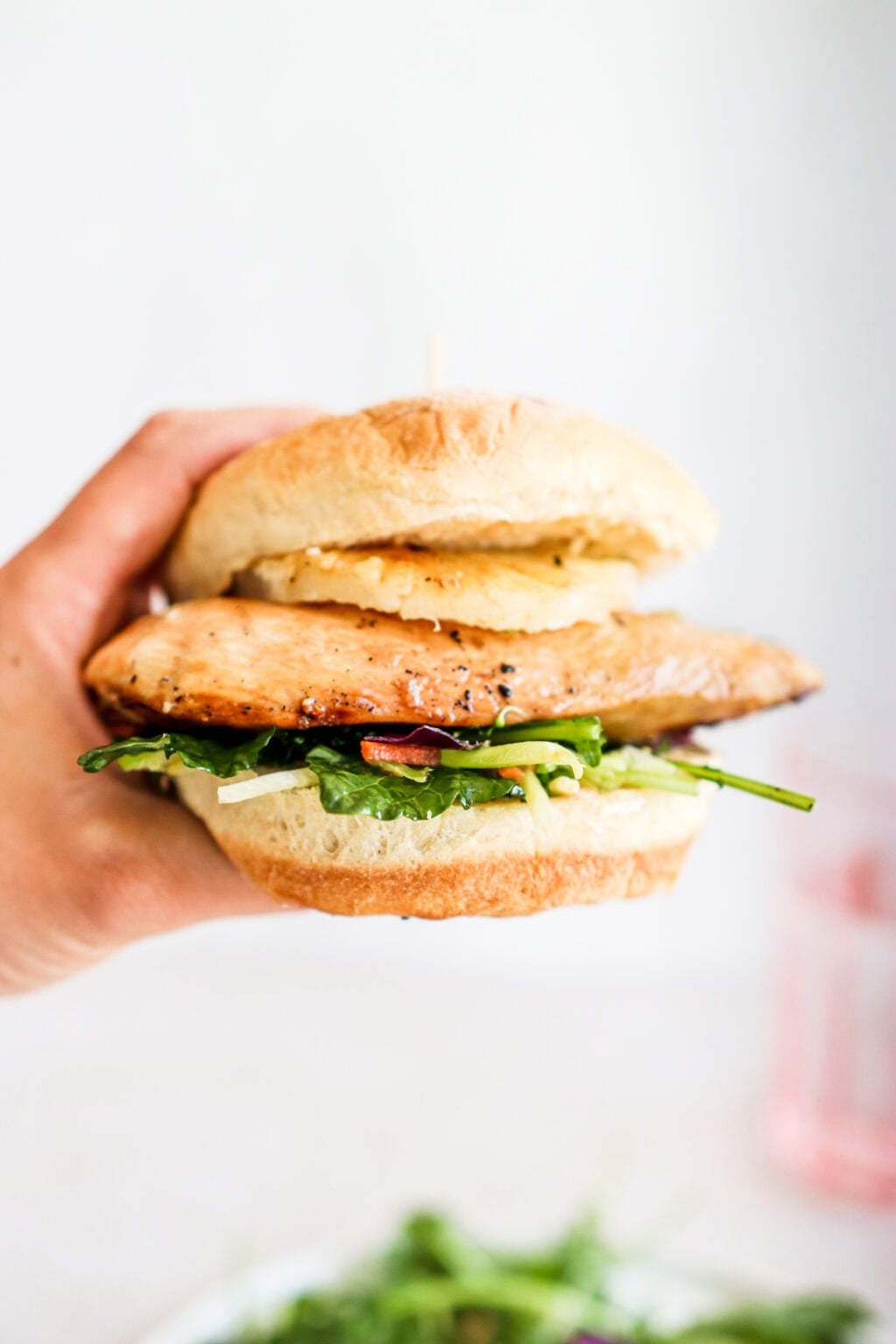 a hand holding a burger bun layered with greens, slaw mix, maple soy glazed chicken breast and a pineapple ring with a pink glass in the background
