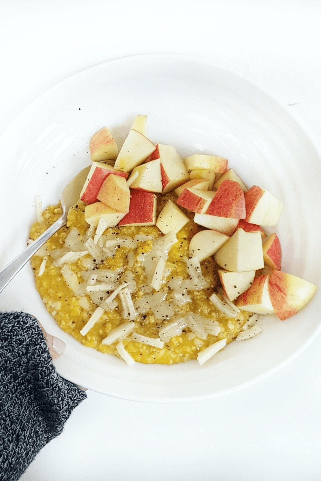 A hand slightly covered by a black sleeve is holding up a white bowl. In the bowl are golden oatmeal topped with diced apple and shredded white cheddar