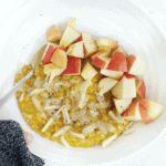 A hand slightly covered by a black sleeve is holding up a white bowl. In the bowl are golden oatmeal topped with diced apple and shredded white cheddar