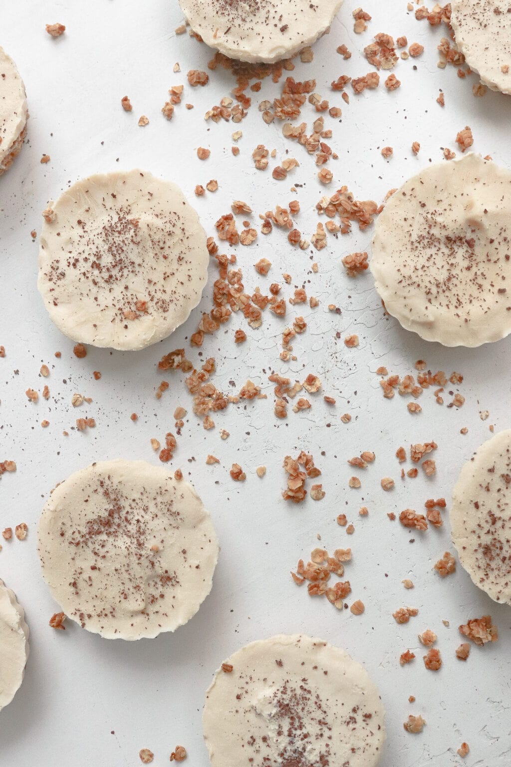 A flat lay photo of 8 cheesecake tarts topped with shaved chocolate. The tarts are surrounded by granola crumble.