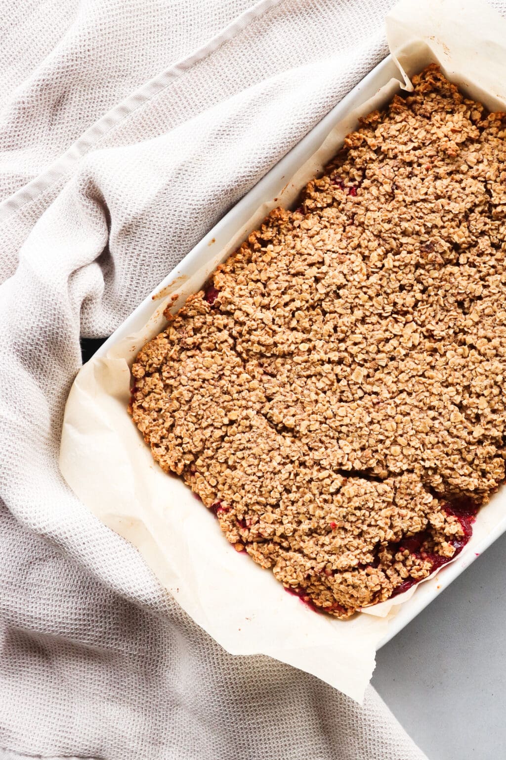 A 9 x 13 inch white baking dish contains baked cranberry oatmeal bars with cranberry sauce. The baking dish is surrounded by an off-white dish towel