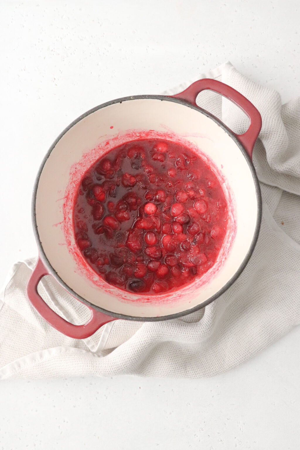 A pot of homemade cranberry sauce is in a red sauce pant and surrounded by a dish towel