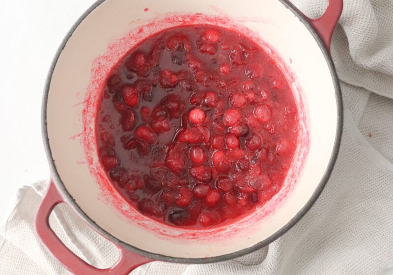 A pot of homemade cranberry sauce is in a red sauce pant and surrounded by a dish towel