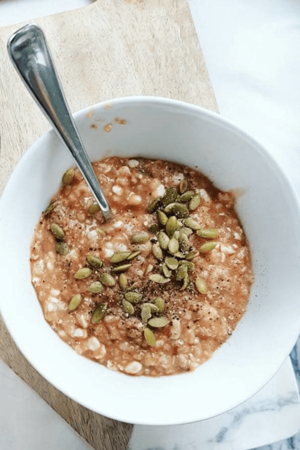 A bowl of stovetop pizza oatmeal topped with pumpkin seeds is sitting on a wooden cutting board. There is a silver spoon in the bowl of oats.