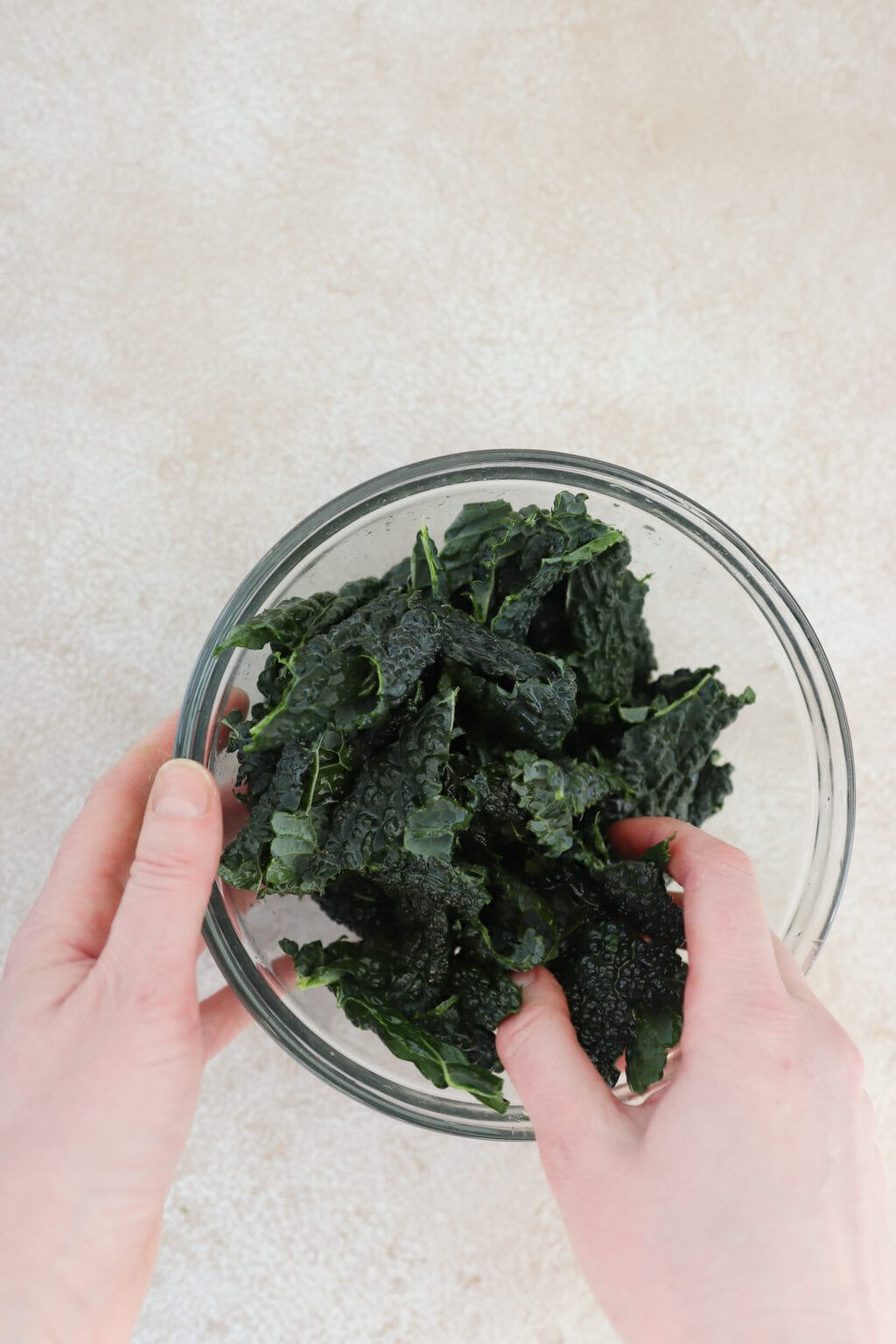 one hand is holding a medium clear bowl, the other hand is massaging dino kale that has been torn into bite sized pieces